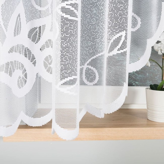 Biała firana gotowa Ewelina wykonana z żakardowego materiału o kwiatowym wzorze, wisi frontalnie na oknie. Obrzeża posiadają falbany. W tle widoczna doniczka z rośliną. Produkt ma wymiary 300x160 cm.