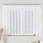 Firana gotowa Felicja żakardowa w kolorze białym, w rozmiarze 400x160 cm, z delikatnym, ażurowym wzorem na dolnej krawędzi. Produkt zawieszony na karniszu, widok frontalny, w jasnym, minimalistycznym wnętrzu, z roślinnością obok.