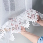 Firana żakardowa, biała, 400x160 cm, z delikatnym, roślinnym wzorem. Ujęcie z bliska, ukazujące rękę podtrzymującą dolny brzeg firany. Tło proste, jasne. Faktura materiału lekka i przezroczysta, z wyraźnym haftem na krawędzi.