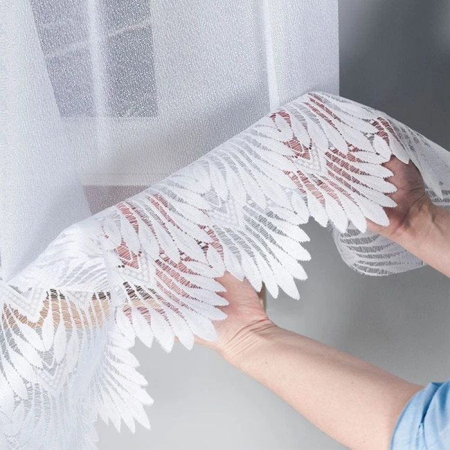 Firana żakardowa, biała, 400x160 cm, z delikatnym, roślinnym wzorem. Ujęcie z bliska, ukazujące rękę podtrzymującą dolny brzeg firany. Tło proste, jasne. Faktura materiału lekka i przezroczysta, z wyraźnym haftem na krawędzi.