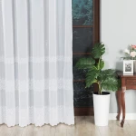 Firana haftowana, biała, z wzorem pasowym, o wysokości 280 cm. Wisząca na oknie, prezentuje subtelne hafty wzdłuż dolnej krawędzi. Otoczenie zawiera ciemną ścianę, roślinę w doniczce i drewniany stolik z dekoracjami.