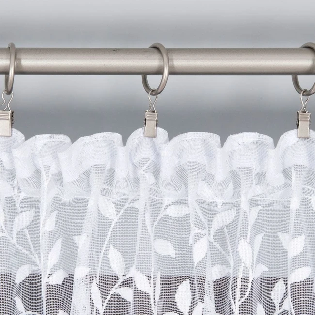 Firana żakardowa Zuzanna w kolorze białym, o wysokości 250 cm, z delikatnym wzorem roślinnym. Wisząca na metalowej karniszu, ukazuje falbany oraz subtelną teksturę materiału. Tło jest jednolite i jasne, co podkreśla elegancję firany.