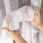 Firana żakardowa w kolorze białym o wysokości 250 cm, z delikatnym wzorem liściowym. Produkt trzymany jest w dłoniach, ukazując jego lekko przezroczystą strukturę oraz falbany na krawędzi. Tło neutralne, co podkreśla detale firany.