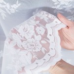 Firana żakardowa w kolorze białym z wzorem kwiatowym na całej powierzchni, o wysokości 250 cm. Produkt wiszący, uchwycony w zbliżeniu, z ręką trzymającą krawędź. Materiał transparentny, teksturowany, wykończony falbanką. Tło jednolite.