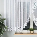 Firana gotowa panelowa Jadwiga żakardowa w kolorze białym, o wymiarach 400x150 cm. Produkt powieszony w oknie, z wyraźnym wzorem w postaci kwiatów na dolnej krawędzi. W tle widoczna roślinność oraz minimalistyczny dekoracyjny element.