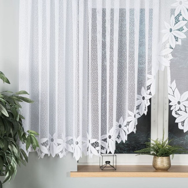 Firana gotowa panelowa Jadwiga żakardowa w kolorze białym, o wymiarach 400x150 cm. Produkt powieszony w oknie, z wyraźnym wzorem w postaci kwiatów na dolnej krawędzi. W tle widoczna roślinność oraz minimalistyczny dekoracyjny element.