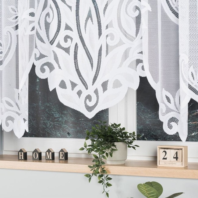 Biała firana żakardowa o wymiarach 500x160 cm, z motywem roślinnym, wisząca w oknie. Tło z zewnątrz przedstawia zieleń, a na parapecie znajdują się ozdobne elementy: doniczka z rośliną i dekoracyjne kostki z napisem HOME oraz datą.