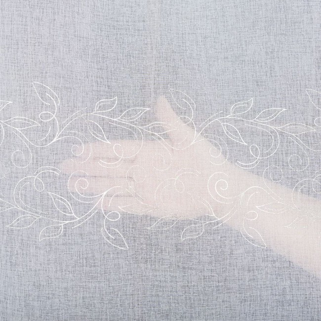 Firana fantazyjna w kolorze białym z delikatnym wzorem ołowianka, wysokość 290 cm. Widoczny wzór roślinny w formie liści oraz skrętów, materiał lekko przezroczysty, uniesiona ręka pokazująca fakturę tkaniny.