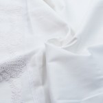 Pościel z gipiurą 160x200 w kolorze białym, wykonana z bawełnianej satyny, leżąca na gładkiej powierzchni. Widoczny detal koronkowej dekoracji wzdłuż brzegu. Faldy tkaniny ukazują gładką fakturę materiału.