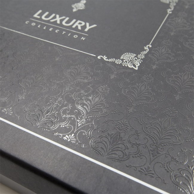 Elegancka pudełko w kolorze ciemnoszarym z wypukłym wzorem roślinnym. Na wieczku widoczny napis LUXURY COLLECTION w srebrnym kolorze. Kąt ujęcia z góry, ukazujący detale faktury i wykończenia pudełka.