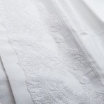 Biała pościel satynowa 220x200 cm z gipiurą, ukazana w zbliżeniu na detal. Widoczny haft w postaci misternych wzorów na jednym z brzegów. Materiał gładki, bez widocznych napisów czy logo. Położona na jednolitym tle, podkreślającym jej fakturę.