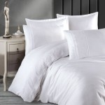 Pościel satyna bawełniana 220x200 Valinor w kolorze białym, z subtelną gipiurą na poszewkach. Ułożona na łóżku w aranżacji sypialni, z widocznymi poduszkami i kołdrą. Tło neutralne, z eleganckim stolikiem nocnym oraz figurką dekoracyjną.