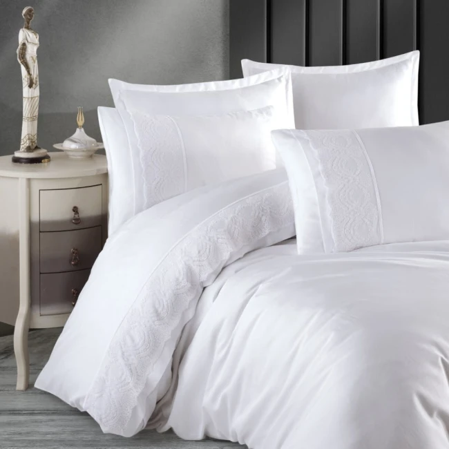 Pościel satyna bawełniana 220x200 Valinor w kolorze białym, z subtelną gipiurą na poszewkach. Ułożona na łóżku w aranżacji sypialni, z widocznymi poduszkami i kołdrą. Tło neutralne, z eleganckim stolikiem nocnym oraz figurką dekoracyjną.