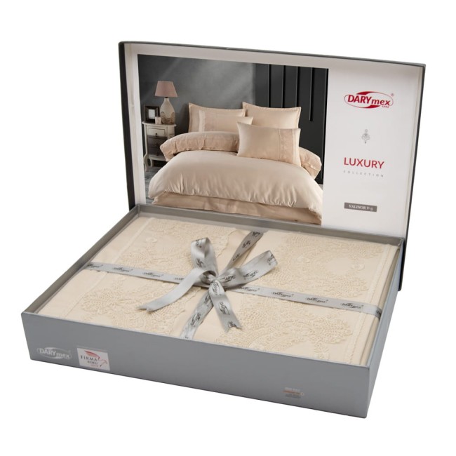 Pościel satyna bawełniana z gipiurą 220x200 Valinor w kolorze beżowym, prezentowana w eleganckim opakowaniu z szarym, satynowym paskiem. Widoczny wzór gipiury na poszwa, rozłożona na tle z aranżacją sypialni. W zestawie dwie poduszki.