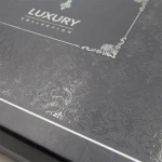 Eleganckie opakowanie w kolorze ciemnoszarym z delikatnym, wypukłym wzorem w stylu roślinnym. Na wieczku widoczny napis „LUXURY COLLECTION” w białej czcionce. Detal w formie subtelnych zdobień dopełnia całość.