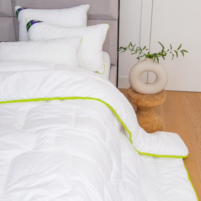 Poduszka antyalergiczna pikowana 50x60 cm Bamboo w białej tkaninie z zielonym obszyciem, leżąca na łóżku obok kołdry. W tle widoczne poduszki o różnych kształtach oraz dekoracyjny wazon z rośliną na okrągłym stoliku.