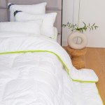 Kołdra całoroczna 200x220 cm w kolorze białym z zielonym brzegi, rozłożona na łóżku, obok poduszki w tym samym kolorze. Widoczne detale pikowania i tekstura materiału. W tle stół z okrągłym wazonem na drewnianej podłodze.
