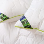 Kołdra całoroczna 200x220 cm, biała, antyalergiczna z materiału Bamboo. Widoczne wzory pikowania oraz zielona lamówka. Ujęcie frontalne, kołdra leży na białym tle, blisko poduszek z logo i etykietą.