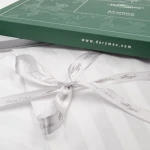 Pościel bambusowa satynowa 160x200 w białe paski, zapakowana w zielone opakowanie z logo DARYmex. W widoku z góry, widoczna satynowa tkanina oraz biała wstążka zdobiona napisem Luxury Collection. Opakowanie ma elegancki, prosty design.