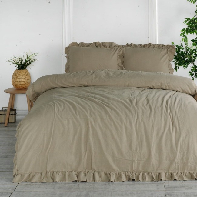 Komplet poszewek bawełnianych z falbanką w odcieniu szaro-beżowym, składający się z dwóch poduszek 40x40 cm. Ułożony na łóżku z materiałem o lekko wrinklowanej fakturze, z ozdobnym falbaniem oraz widocznymi poduszkami. W tle roślinność i drewniane meble.