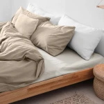 Prześcieradło flanelowe bez gumki w kolorze kremowym 180x210, leżące na łóżku, obok poduszki. Widać teksturę materiału oraz rozłożoną fakturę. Na łóżku znajdują się także białe poduszki oraz wiklinowy kosz w tle.