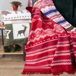Czerwony koc bawełniany o wymiarach 150x200 cm, z białymi i niebieskimi wzorami w świąteczne motywy. Koc z frędzlami, leżący na stole obok białego koca z zielonymi detalami i motywem jelenia. Tło w odcieniach brązu i zieleni.