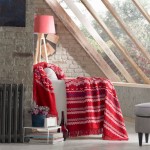 Koc bawełniany w kolorze czerwonym o wymiarach 150x200 cm, z białymi i niebieskimi wzorami w stylu świątecznym. Leży na kanapie w jasnym wnętrzu, obok znajduje się kwiatek w doniczce oraz stos książek.