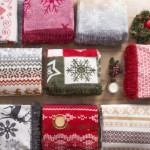 Koc bawełniany 150x200 cm w czerwonym kolorze, z białymi i zielonymi wzorami świątecznymi, umieszczony w zestawie z innymi kocami. Koc ma ozdobne frędzle. W tle znajduje się świeczka i dekoracje bożonarodzeniowe.