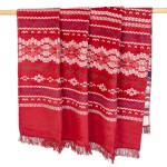 Koc bawełniany 150x200 cm w czerwonym kolorze z białymi, niebieskimi i zielonymi wzorami świątecznymi. Zakończony frędzlami, zawieszony na drewnianym wieszaku. Idealny do dekoracji wnętrz w okresie świątecznym.