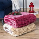 Koc bawełniany 150x200 w bordowym kolorze, z frędzlami na jednym końcu, leży na drewnianej powierzchni obok beżowego koca. W tle widoczna lampa i dekoracje świąteczne. Koc ma delikatny wzór, a jego faktura jest miękka i przyjemna w dotyku.