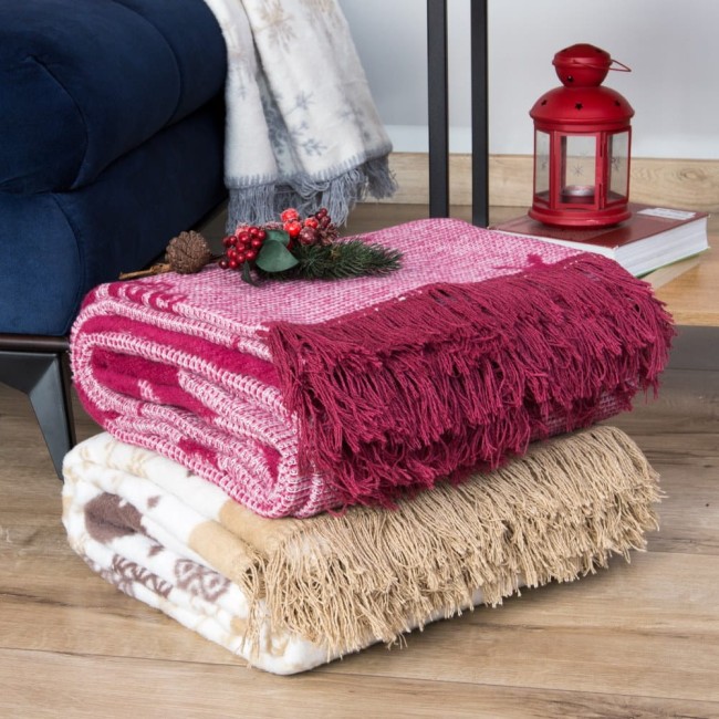 Koc bawełniany 150x200 w bordowym kolorze, z frędzlami na jednym końcu, leży na drewnianej powierzchni obok beżowego koca. W tle widoczna lampa i dekoracje świąteczne. Koc ma delikatny wzór, a jego faktura jest miękka i przyjemna w dotyku.