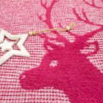 Koc bawełniany 150x200 cm w bordowym kolorze, z wzorem renifera. Produkt leży na płaskiej powierzchni, ujęcie w zbliżeniu. Widoczna tekstura materiału, dekoracyjna gwiazda w białym kolorze obok koca. Tło w harmoni z wzorem, nadające świąteczny charakter.