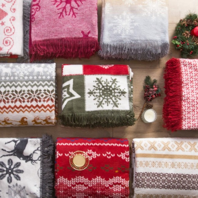 Bordowy koc bawełniany 150x200 cm z frędzlami, zdobiony świątecznym wzorem w gwiazdy i płatki śniegu. Produkt leży na drewnianej powierzchni, otoczony innymi kocami w różnych kolorach i wzorach, z dekoracyjnymi akcentami w tle.