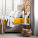 Prześcieradło frotte z gumką w kolorze żółtym, rozłożone na materacu, z widoczną teksturą materiału. Obok poduszki w jasnym kolorze i dekoracyjne elementy na stoliku. Tło stanowi elegancki wzór na ścianie oraz lekko rozświetlone zasłony.