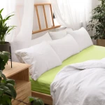 Prześcieradło jersey z gumką w kolorze limonkowym, o wymiarach 90x200 cm, umieszczone na łóżku z białymi poduszkami. W tle widoczne rośliny oraz elementy drewniane, co tworzy przytulną atmosferę.