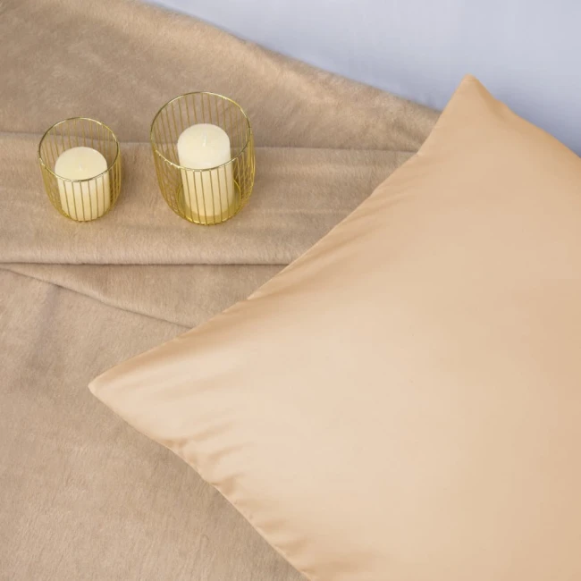Poszewka satynowa w kolorze jasnego beżu, o wymiarach 70x80 cm, leży na gładkim tle. Ujęcie z góry pokazuje jej gładką fakturę. Obok znajdują się dwa szklane świeczniki z białymi świecami, tworząc aranżację.