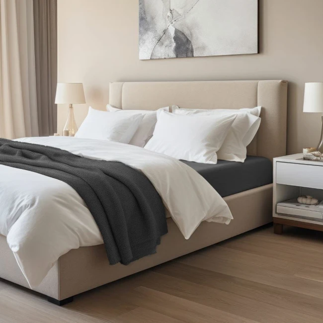 Prześcieradło jersey z elastanem z gumką w kolorze czarnym, rozłożone na materacu o wymiarach 180x200 cm. Ujęcie z boku, ukazuje również białą pościel i szary pled na łóżku w nowoczesnej sypialni.