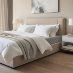 Prześcieradło jersey z elastanem w kolorze popiel z gumką, dostępne w rozmiarze 180x200, leży na materacu, otoczone białymi poduszkami i narzutą w neutralnych odcieniach, w nowoczesnej sypialni z jasnym tłem i naturalnym drewnem.