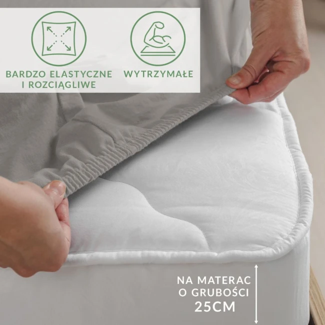 Prześcieradło jersey w kolorze popielatym, z gumką, rozłożone na białym materacu. Ujęcie z boku, pokazujące elastyczność materiału oraz mocowanie. Widoczny napis informujący o dopasowaniu do materaca o grubości 25 cm.
