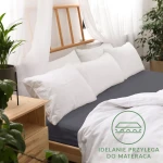 Prześcieradło jersey z gumką 90x200 w kolorze antracytowym leży na łóżku, otoczone białymi poduszkami. Widoczna faktura materiału, dopasowujące się do materaca krawędzie, dekoracyjna etykieta z napisem idealnie przylega do materaca.