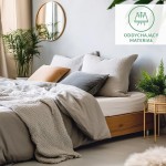 Flanelowe prześcieradło z gumką w kolorze jasno szarym, rozłożone na łóżku z drewnianym stelażem. W tle widoczne poduszki w odcieniach szarości i pomarańczu, roślinność oraz okrągłe lustro. Tekstura materiału matowa i delikatna.