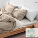 Prześcieradło flanelowe z gumką w kolorze kremowym, rozmiar 200x220 cm, leżące na łóżku z poduszkami. Widoczna tekstura materiału, zapewniającego komfort. Na białym tle, bez dodatkowych dekoracji, z logo „nie zsuwają się z materaca” w rogu.