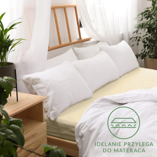 Prześcieradło jersey z gumką w kolorze kremowym, rozmiar 160x200 cm, rozłożone na łóżku, idealnie przylegające do materaca. W tle widoczne białe poduszki i drewniane łóżko. Tło jasne, o naturalnej aranżacji. Logo z napisem widoczne w dolnym rogu.