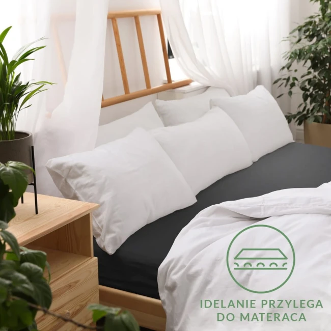 Prześcieradło jersey z gumką w kolorze czarnym, rozmiar 140x200, leży na łóżku z białymi poduszkami. Ujęcie z góry, ukazujące detail фактуры tkaniny. W tle widoczne drewniane elementy i roślinność, całość w jasnym, naturalnym otoczeniu.