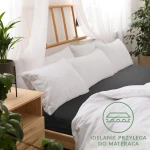 Prześcieradło jersey z gumką 90x200 w kolorze czarnym, znajdujące się na łóżku z białymi poduszkami. Produkt rozłożony na materacu, z widocznymi detalami gumki, dobrze przylega do tkaniny. Tło zawiera drewniane meble i rośliny w donicach.