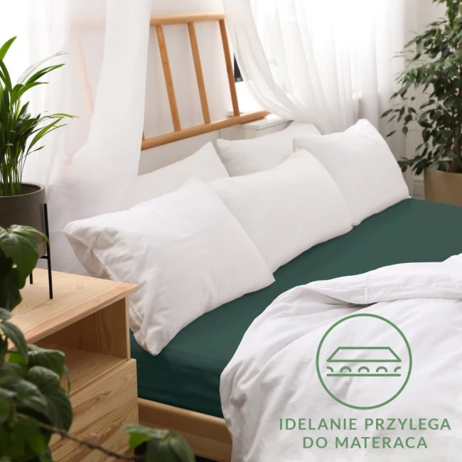 Prześcieradło jersey z gumką 120x200 w kolorze butelkowej zieleni, rozłożone na łóżku z białą pościelą. Widoczna guma brzegowa. W tle drewniane elementy, roślinność i okno z zasłonami. Elegancka aranżacja sypialni, zbliżenie na materiał.