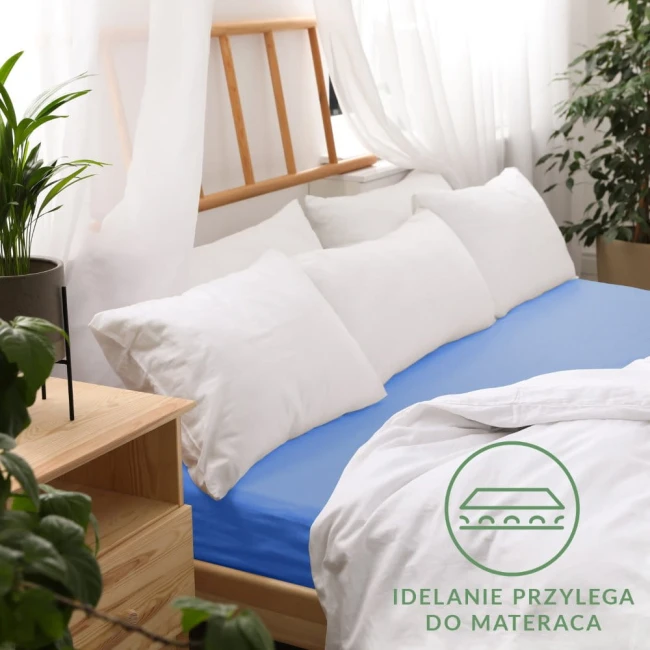 Prześcieradło jersey z gumką 180x200 w kolorze chaber, rozłożone na łóżku z białymi poduszkami. Kąt ujęcia frontalny, ukazujący sypialnię z naturalnym światłem. Widoczna informacja Idealnie przylega do materaca. Tło jasne, z drewnianymi elementami.