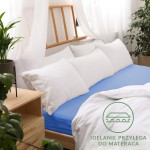 Prześcieradło jersey z gumką 200x220 w kolorze chabrowym, leżące na łóżku z białymi poduszkami. Materiał gładki, dobrze przylegający do materaca. Tło stanowi jasne wnętrze z naturalnymi akcentami roślinnymi. Widoczne detale, jak elastyczna gumka.