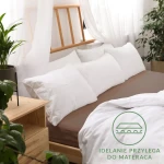 Prześcieradło jersey z gumką w kolorze czekoladowym leży na łóżku, w zestawieniu z białymi poduszkami. Widoczna tekstura materiału i gumka, która idealnie przylega do materaca. Tło to jasne wnętrze z drewnianym wezgłowiem i roślinnością.