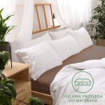 Prześcieradło jersey z gumką 140x200 w kolorze czekoladowym, leżące na łóżku. Na prześcieradle widoczne białe poduszki i część białej pościeli. Tło jasne z drewnianą ramą łóżka i roślinnością. Widoczny napis Idealnie przylega do materaca.